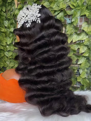 Long, dark brown to black wavy hair extension or wig displayed on a mannequin head, styled with a white floral headpiece, set against a green leaf wall background.