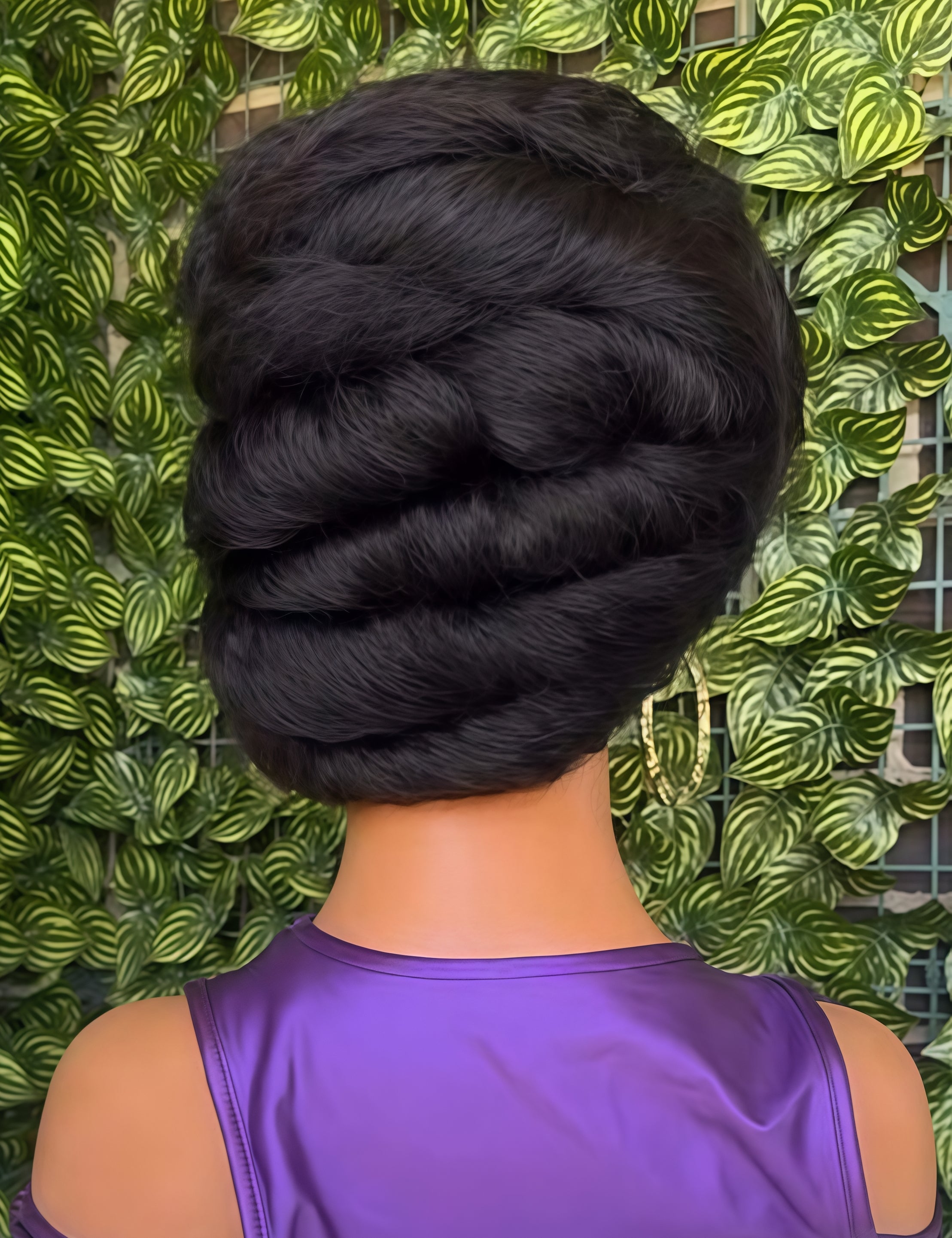 A close-up back view of a mannequin or dress form wearing a shiny purple top, with a voluminous black braided hairstyle. The background is a dense green leafy wall.