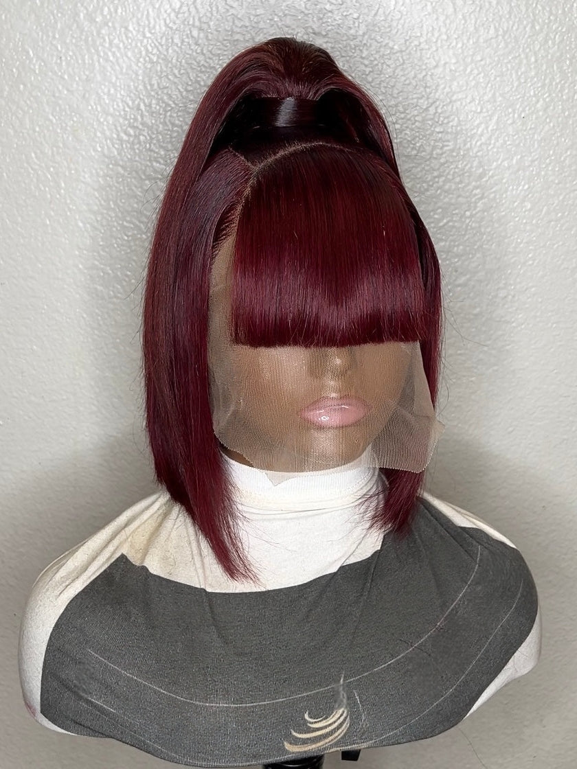 A short, straight wig with deep burgundy highlights and dark roots, styled on a mannequin bust with a white turtleneck and gray bib against a textured white wall.