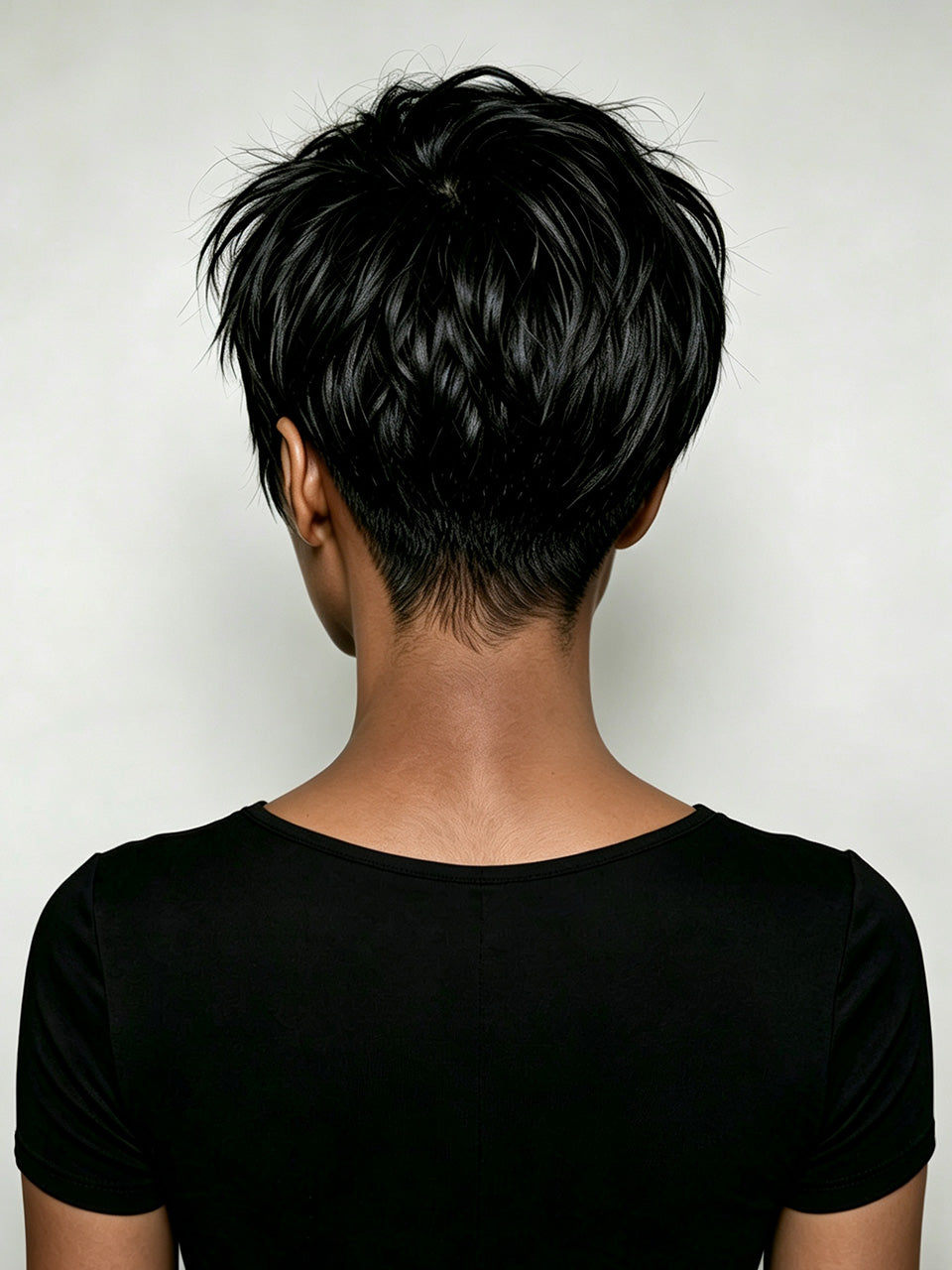 Rear view of a person with a short, layered black pixie cut, wearing a black short-sleeve top against a light gray background.