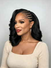 A woman with long, black, wavy hair styled with cornrow braids along the side, wearing a cream ribbed long-sleeve top against a light gray background.