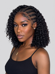 A woman with a short, curly black wig styled with side-parted braids, wearing a black ribbed tank top against a solid light gray background.