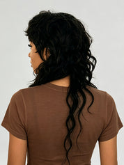A woman shown from the back wearing a brown short-sleeve t-shirt and a long, wavy black wig. The wig is the primary product being showcased here, displayed on a model against a plain light gray background.