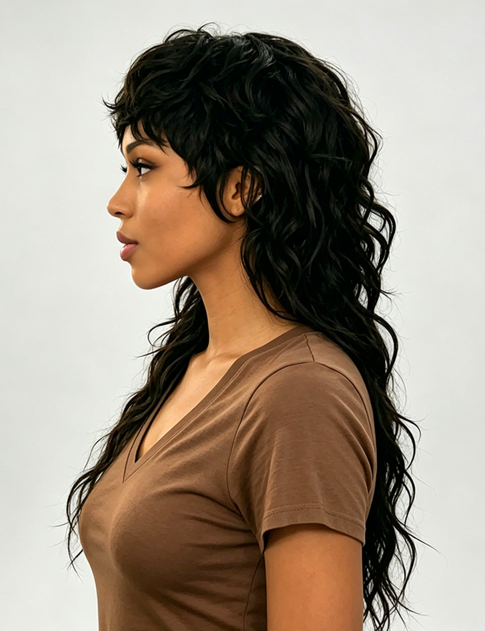A woman shown in side profile wearing a long, dark brown to black wavy wig with layered bangs and a side-swept look, paired with a brown short-sleeve V-neck top against a light gray studio background.