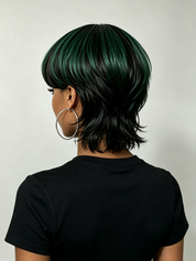 Rear view of a person wearing a short layered wig with dark black and deep emerald green streaks, paired with a black t-shirt and large silver hoop earrings against a light gray background.
