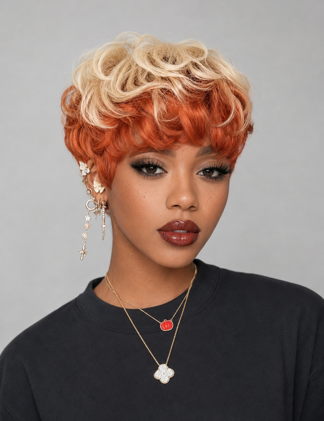 A woman wearing a short, curly wig with a blonde-to-orange ombre color transition, paired with gold-toned jewelry and a black top, against a plain gray background.