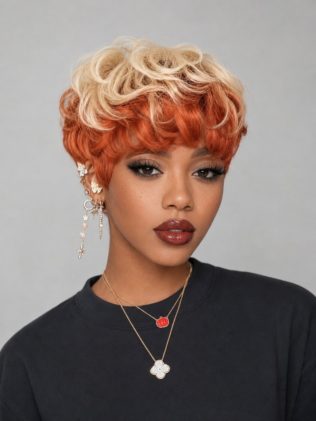 A woman wearing a short, curly wig with a blonde-to-orange ombre color transition, paired with gold-toned jewelry and a black top, against a plain gray background.