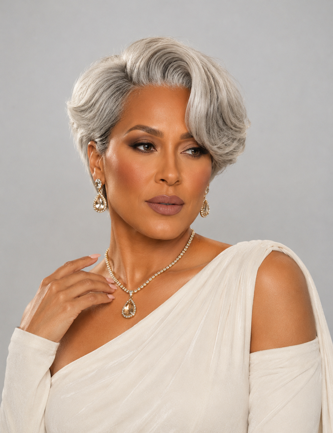A woman with a short, styled silver-gray wig, wearing a white draped off-the-shoulder top and gold jewelry, posing against a solid gray studio background.