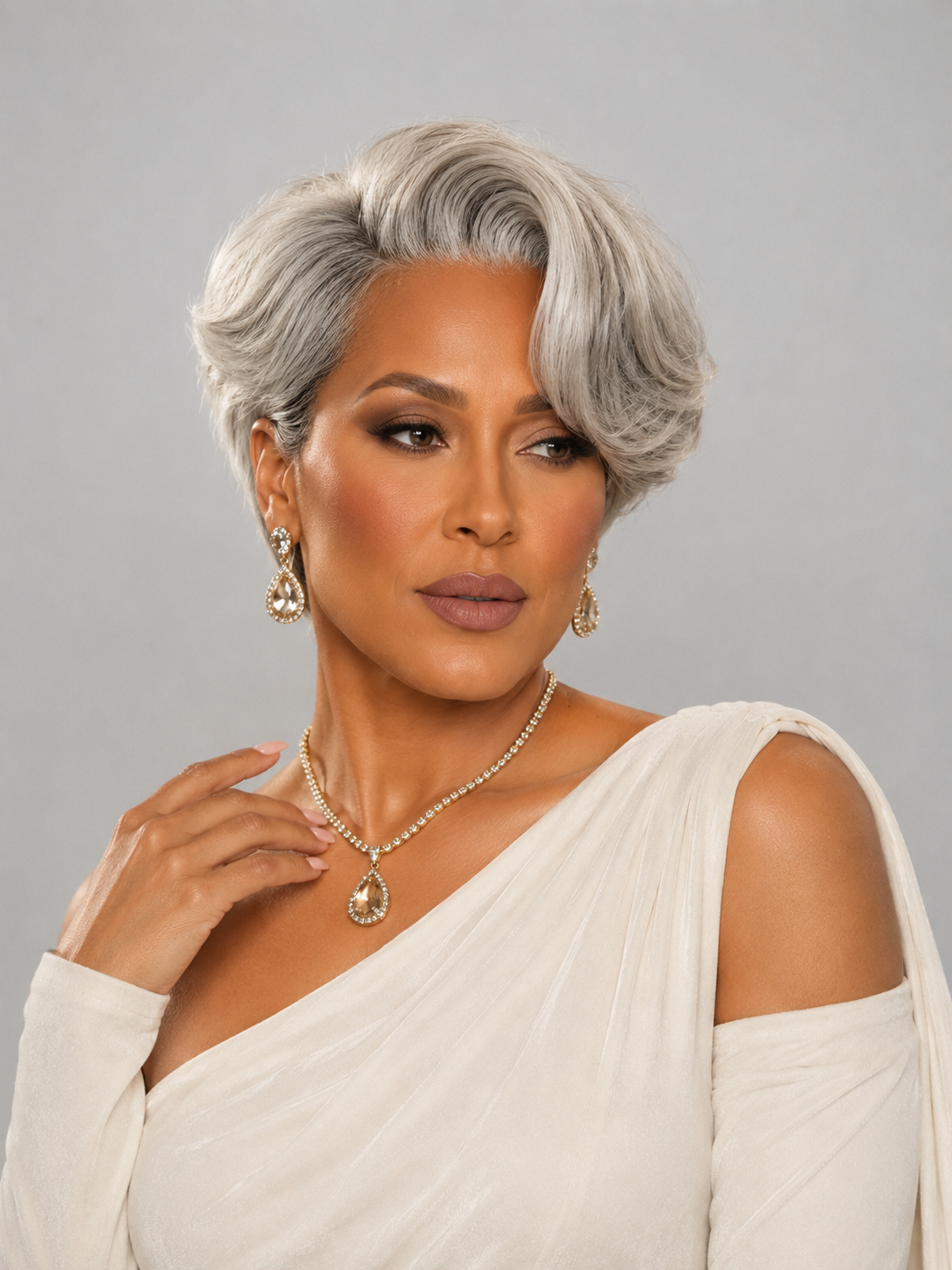 A woman with a short, styled silver-gray wig, wearing a white draped off-the-shoulder top and gold jewelry, posing against a solid gray studio background.