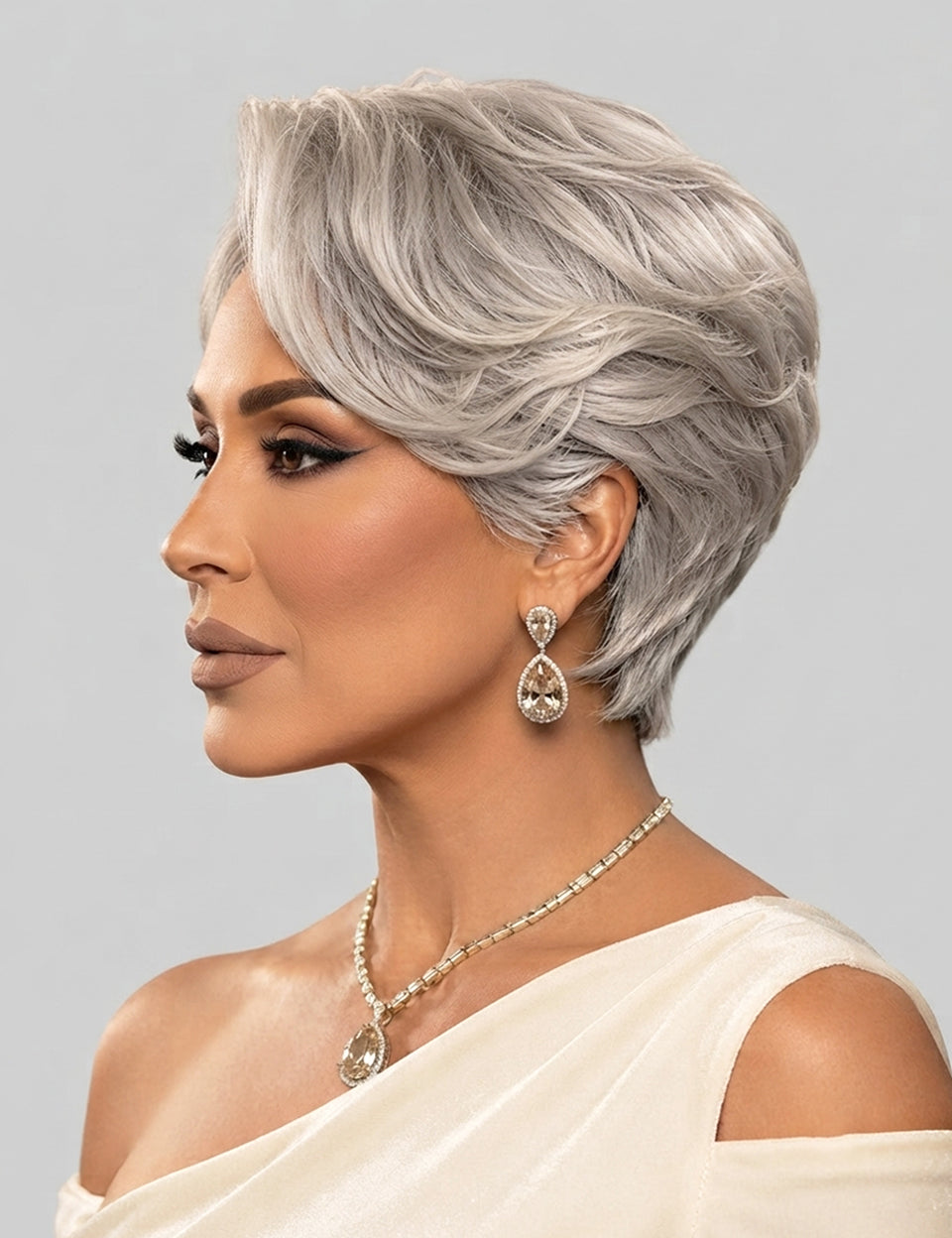 A woman shown in profile wearing a short, layered silver-gray wig, along with a matching silver teardrop earring and necklace, against a light gray studio background.