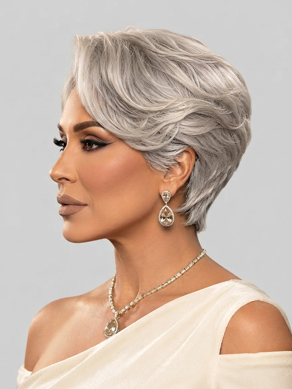 A woman shown in profile wearing a short, layered silver-gray wig, along with a matching silver teardrop earring and necklace, against a light gray studio background.
