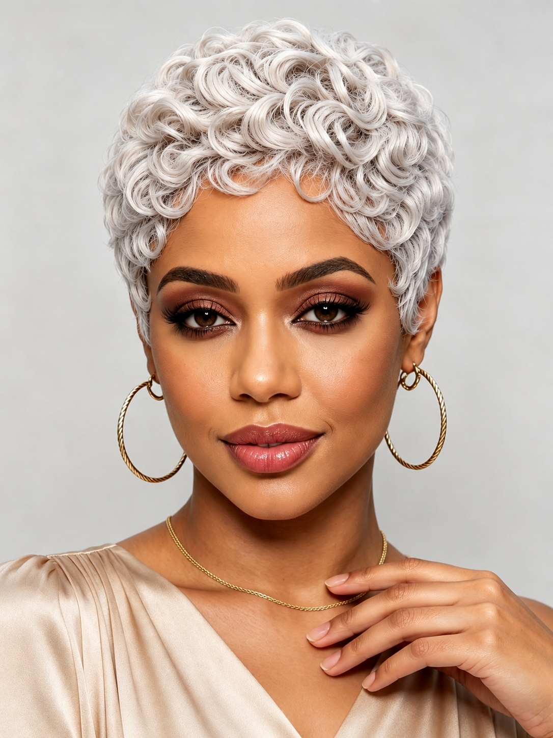 A woman with a short, curly platinum-blonde wig, gold hoop earrings, and a gold necklace, wearing a champagne-colored satin top against a light gray background.