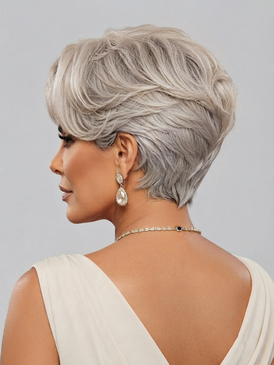 A woman shown from the back and side profile wearing a short, layered silver-gray wig, a delicate gold chain necklace, and dangling pearl-like earrings against a light gray studio background.