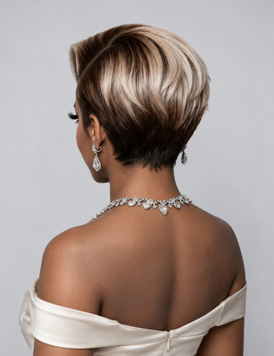 A woman shown from the back wearing a short layered haircut with dark brown roots and blonde highlights, styled with a silver jeweled necklace and earrings against a light gray background.