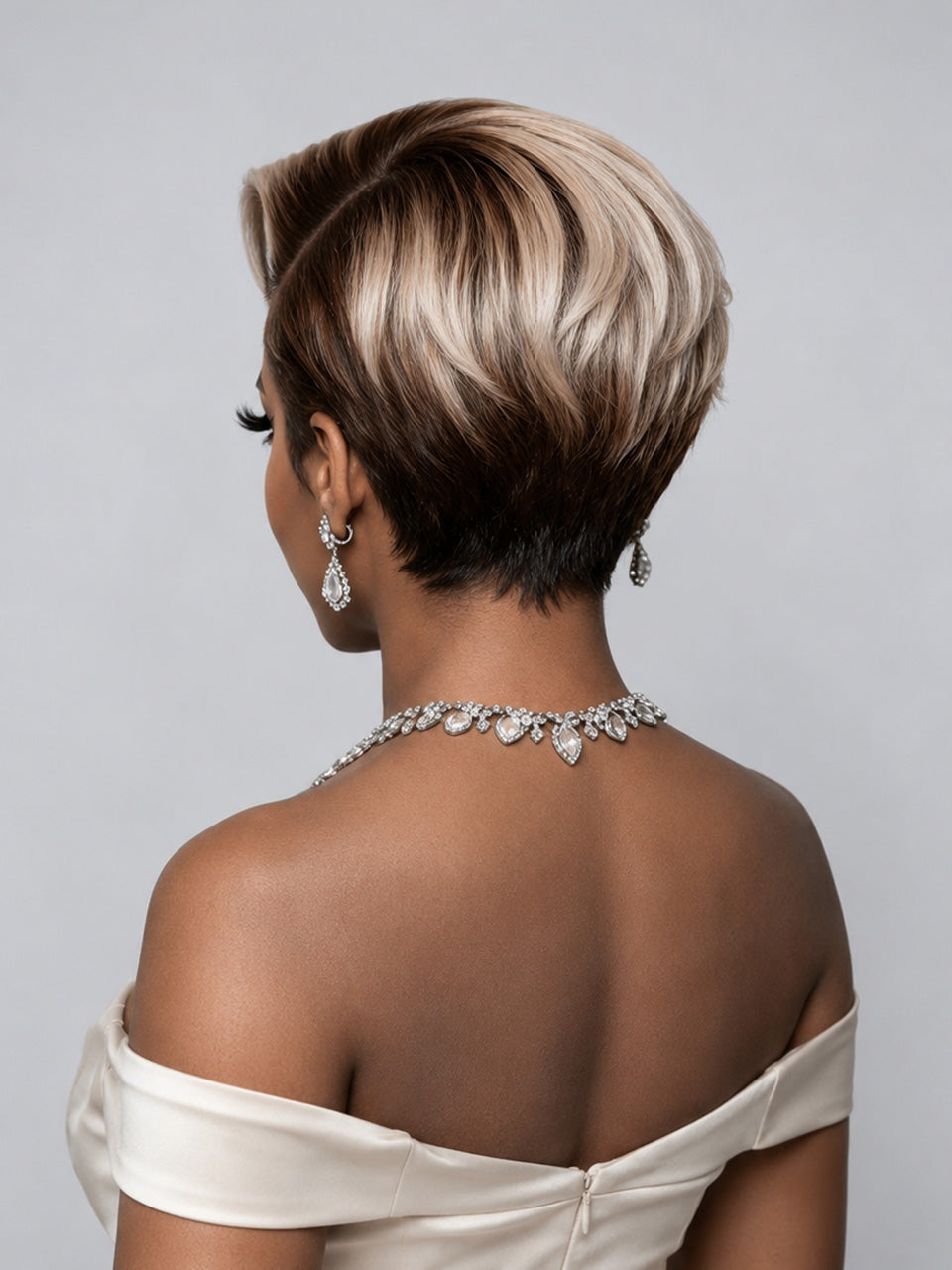 A woman shown from the back wearing a short layered haircut with dark brown roots and blonde highlights, styled with a silver jeweled necklace and earrings against a light gray background.
