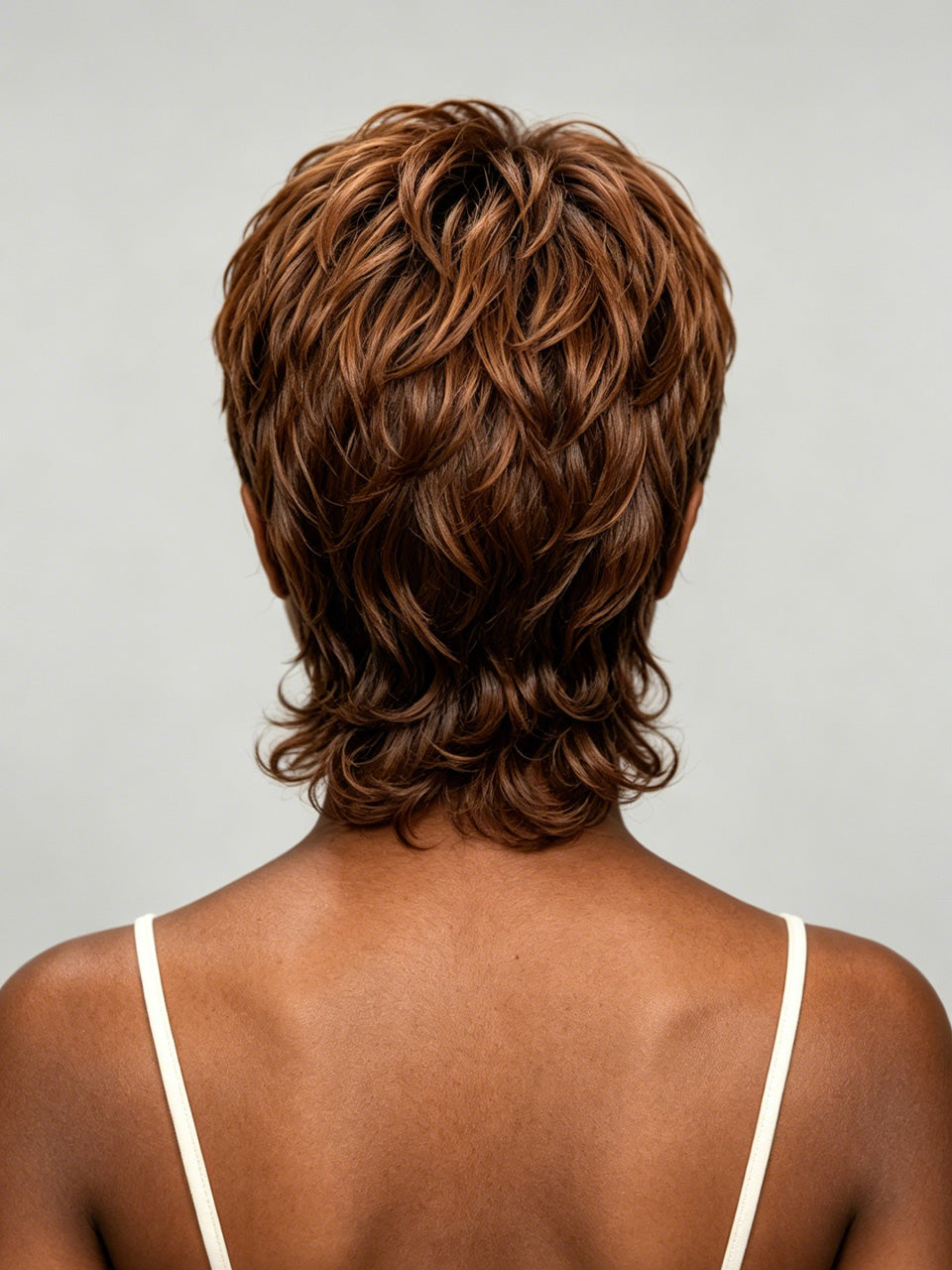 Rear view of a person with a short, layered, wavy wig in medium-to-dark brown with warm caramel highlights, shown against a light gray studio background.