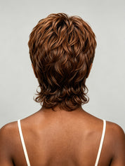 Rear view of a person with a short, layered, wavy wig in medium-to-dark brown with warm caramel highlights, shown against a light gray studio background.