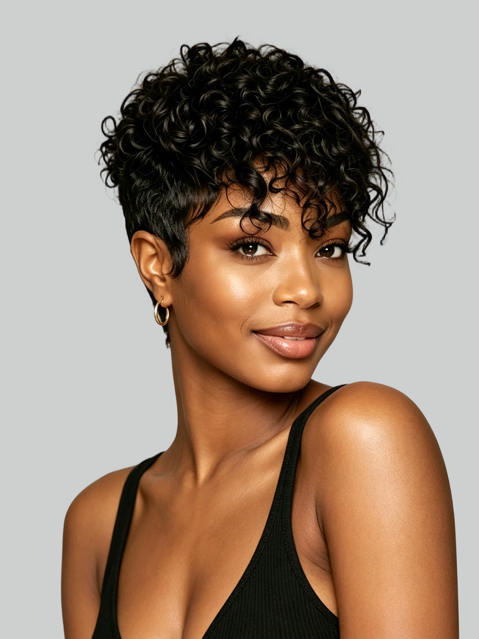A woman with short, curly black hair wearing a black ribbed tank top against a solid gray studio background.