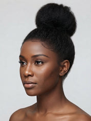 A close-up portrait of a woman with a high, voluminous black hair bun styled in a natural afro texture, shown against a plain light gray background.